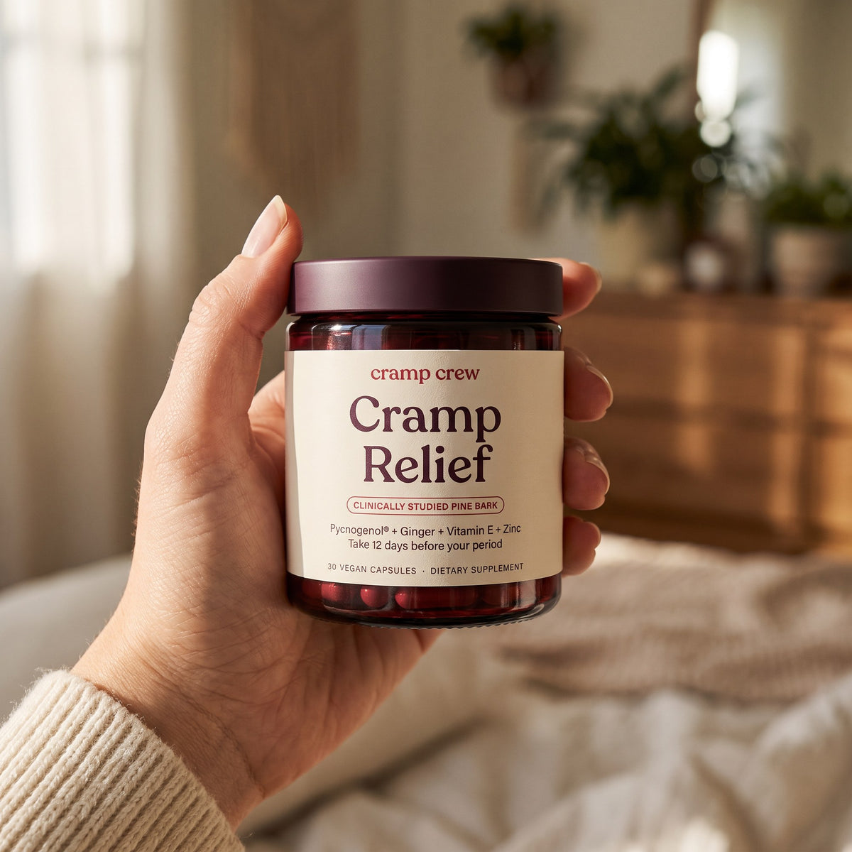 Hand holding a jar of cramp crew Cramp Relief in a cosy bedroom