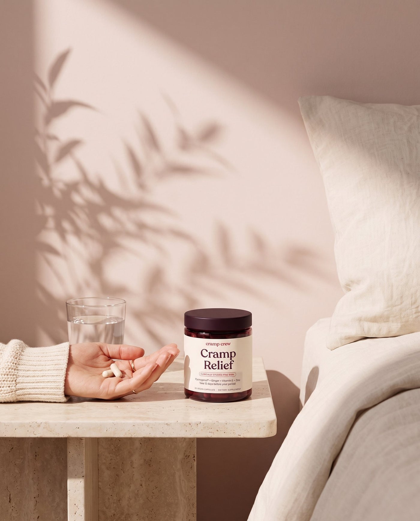 A hand holding two Cramp Relief capsules next to the jar on a bedside table